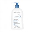 Un flacon blanc de 1 litre de Bioderma Atoderm Crème de Douche avec une pompe bleue. L'étiquette précise qu'il est ultra-nourrissant, adapté aux peaux sensibles normales à sèches, et mentionne « Laboratoire Dermatologique ». La bouteille se reflète partiellement sur une surface blanche.
