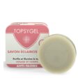 L'image montre une barre ronde de savon Topsygel devant son emballage rectangulaire. L'emballage est majoritairement rose et blanc et porte une étiquette en français, indiquant qu'il s'agit d'un savon éclaircissant concentré pour le visage et le corps, conçu pour purifier et illuminer la peau.