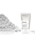 Un tube de produit de soin pour la peau The Ordinary Vitamin C Suspension 23 % + HA Spheres 2 % se tient debout à côté d'un tas de poudre blanche. Le tube est blanc avec un texte minimaliste en noir et gris détaillant le nom et la formulation du produit. Le fond est blanc uni.