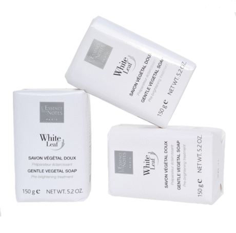 Trois barres rectangulaires de Savon Végétal Doux Feuille Blanche sont exposées. Chaque barre est emballée dans un emballage blanc avec du texte en noir et gris. Le texte comprend "L'Essence des Notes", "Paris", ainsi que des indications sur le poids du savon (150 g) et ses bienfaits (soin pré-éclaircissant).
