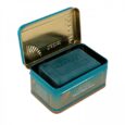 Une boîte rectangulaire ouverte en métal bleu sarcelle et doré contient une barre de savon rectangulaire gris foncé à l'intérieur. Le couvercle est articulé et maintenu ouvert, montrant le nom de marque « MAKARI » en relief à l'intérieur du couvercle et imprimé sur le savon. La boîte comporte également des motifs de texte et de coquillages.