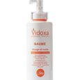 Une bouteille blanche de Vidoza Baume avec un distributeur à pompe. Le produit est destiné au visage et au corps et est commercialisé pour nourrir et hydrater les peaux sèches. Le texte est principalement en français et la bouteille présente des accents et une marque orange.