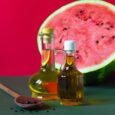 Deux bouteilles d'huile en verre, l'une dotée d'un bouchon en liège en bois et l'autre d'un couvercle blanc, sont placées devant une tranche de pastèque révélant sa chair rouge et ses graines. Une cuillère en bois avec des graines noires repose sur la surface, le tout sur un fond rouge et vert.