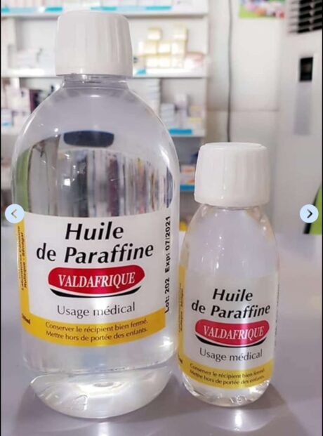 Deux bouteilles en plastique d'Huile de Paraffine avec bouchons blancs et étiquettes jaunes sont placées sur un comptoir. La plus grande bouteille est à gauche, tandis que la plus petite bouteille est à droite. Les deux étiquettes indiquent « Valdafrique » et indiquent un usage médical.