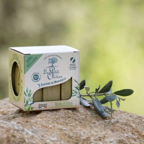 L'image montre une boîte de savons à l'huile d'olive "Le Petit Olivier" posée sur une surface en pierre avec une branche d'olivier à côté. L'emballage est respectueux de l'environnement, soulignant qu'il est composé à 72 % d'huile d'olive et qu'il s'agit d'un savon de Marseille extra doux et naturel.