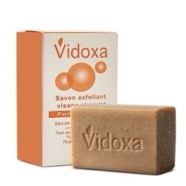 Un pain de savon exfoliant visage marron sur lequel est gravé « VIDOXA Savon 200g » est placé devant son emballage. La boîte est blanche avec des accents orange et porte le nom de marque « VIDOXA Savon 200g », avec un texte en français le décrivant comme un savon exfoliant pour le visage pour purifier la peau.