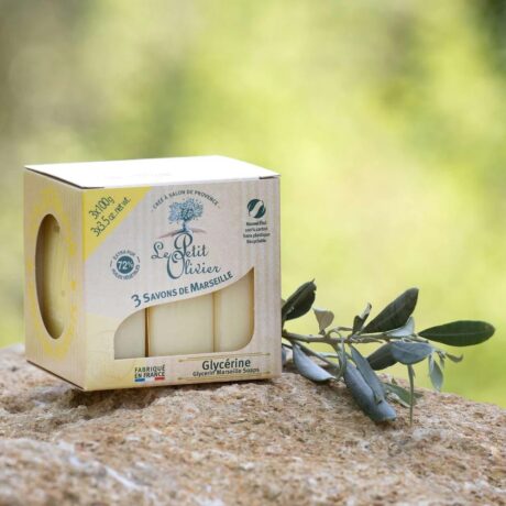 une boîte de savon le petit olivier repose sur une surface en pierre à côté d'une petite branche aux feuilles vertes. l'emballage souligne que le savon est fabriqué en france et contient de la glycérine. l'arrière plan est une verdure floue.