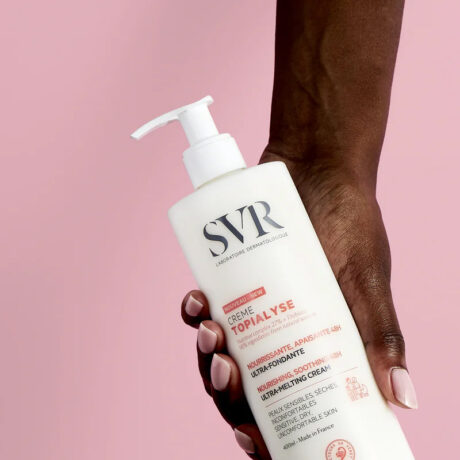 une main tient un flacon de crème topialyse svr sur fond rose. la crème est étiquetée comme nourrissante et apaisante pour les peaux sèches et sensibles, avec un flacon pompe sur le dessus.