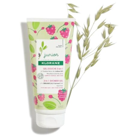 un tube blanc de gel douche 2 en 1 klorane junior au design ludique à base de fruits, avec des fraises et des feuilles vertes. le tube est posé à la verticale à côté d'un brin d'herbe séchée.