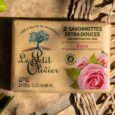 emballage des savons extra doux le petit olivier au parfum de rose, avec un logo d'arbre, des images de roses roses et un texte indiquant qu'il contient deux savons de 100 g. placé sur une surface texturée avec un sac à ficelle à proximité.