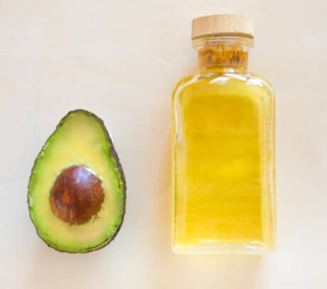 À gauche, un avocat coupé en deux, le noyau exposé. À droite, une bouteille transparente remplie d'huile jaune et fermée par un bouchon en bois. le fond est d'une couleur claire et neutre.