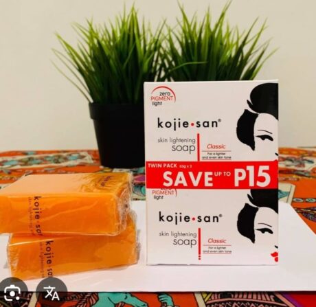 deux barres de savon éclaircissant orange kojie san sont empilées à côté d'une boîte de savon blanche et noire. une plante verte en pot trône en arrière plan sur une table recouverte d'une nappe à motifs colorés.