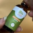une main tient une petite bouteille brune étiquetée « bio nora collagen plus serum » avec une étiquette verte et blanche sur un fond beige.
