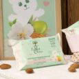 un paquet de lingettes nettoyantes pour bébés le petit olivier est posé sur une surface en bois à proximité d'amandes, de serviettes roses et d'une illustration d'ours encadrée avec des fleurs en arrière plan. l'emballage est vert pastel et blanc avec des accents floraux.