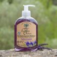 une bouteille de savon liquide le petit olivier parfumé à la lavande avec une pompe, placée sur une surface à l'extérieur. la bouteille transparente contient du savon violet et une étiquette représentant des fleurs de lavande et un arbre. des brins de lavande sont posés devant.