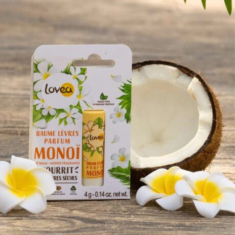 un emballage de baume à lèvres lovea monoi se tient debout à côté d'une noix de coco coupée en deux et de deux fleurs de plumeria blanches et jaunes sur une surface en bois naturel. la scène évoque des ingrédients tropicaux et nourrissants.