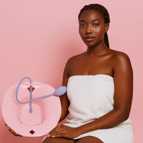 une femme portant une serviette blanche est assise sur un fond rose, tenant un bain de siège rose avec une pompe à main violette et un tuyau d'arrosage.