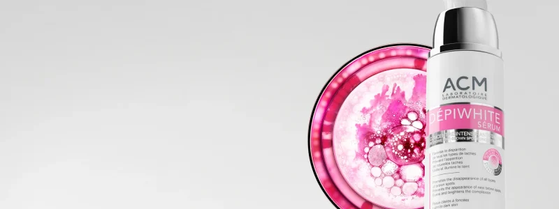 un flacon d'acm depiwhite serum se trouve à côté d'un dessin abstrait circulaire rose et blanc ressemblant à un gros plan de bulles, sur un fond gris clair uni.