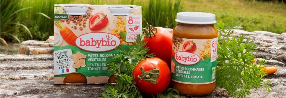 une boîte et un pot de pâtes bolognaises aux légumes babybio avec lentilles et tomates sont posés sur une surface en pierre en plein air, à côté de tomates et de carottes fraîches, avec de la verdure en arrière plan.