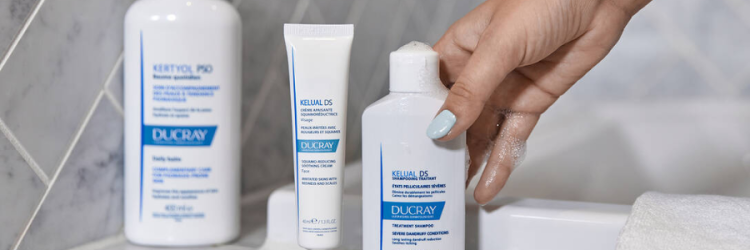 une main aux ongles peints en bleu clair tient une bouteille de shampoing ducray kerium ds sur un comptoir de salle de bain, à côté d'autres produits de soins capillaires ducray et d'une serviette blanche.